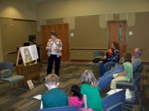 woman presenting oversize book on easel in front of small group of adults and children